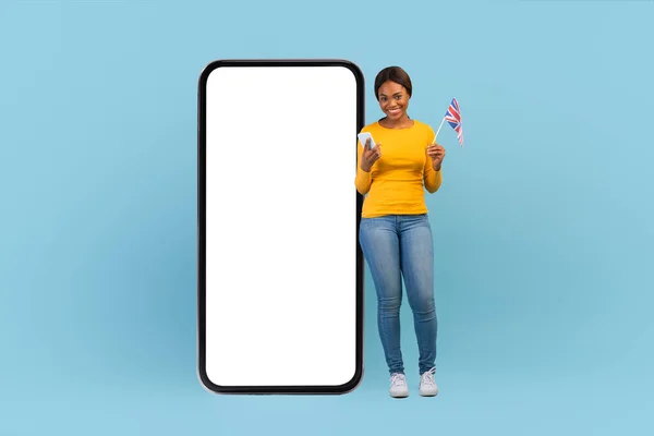 해피 밀레니엄 아프리카 계 미국인 여성, 전화기로 캐묻고, 영국 국기를 들고, 큰 스마트폰을 들고 서서 — 스톡 사진