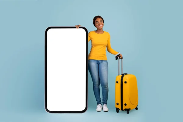Feliz joven hembra negra con billete de avión y pasaporte con maleta se levanta con enorme teléfono inteligente con pantalla en blanco — Foto de Stock