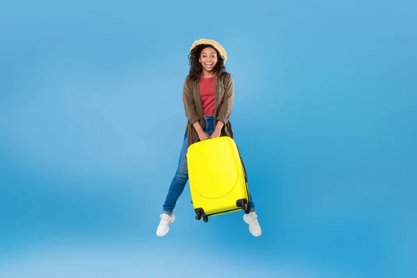 Aufgeregte afroamerikanische Millennial-Frau in lässigem Outfit mit aufspringendem Koffer in der Luft auf blauem Hintergrund — Stockfoto