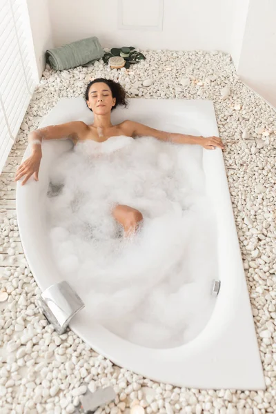 Wanita muda yang cantik berbaring di bak mandi dengan mata tertutup, menikmati mandi santai di spa mewah, pemandangan atas — Stok Foto
