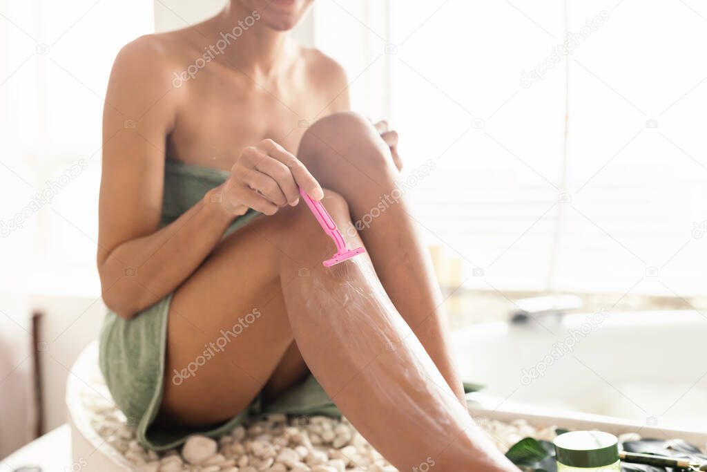 Cropped view of young woman shaving legs with razor, removing unwanted body hair, making epilation near bubble bath