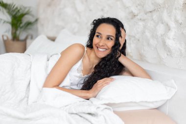 Tatmin olmuş beyaz saçlı, uzun kıvırcık saçlı, pijamalı bir kadın. Boş zamanların tadını çıkarıyor. Yatakta uzanıyor.