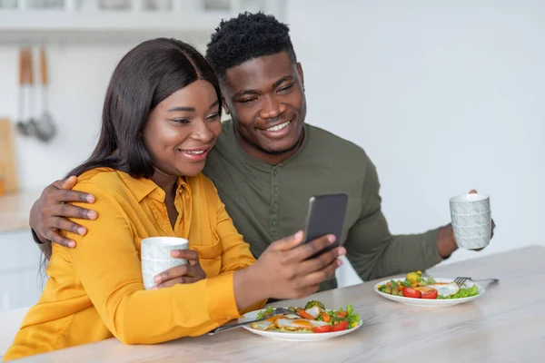 Jeune homme noir et femme en utilisant Smartphone tout en appréciant le petit déjeuner dans la cuisine — Photo