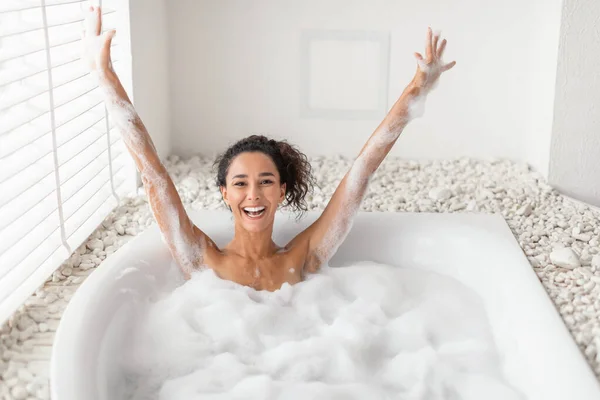 Excited millennial woman taking bubble bath, raising arms up, having fun on relaxing weekend at home, copy space — ストック写真