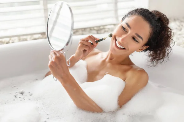 Adorável jovem mulher fazendo massagem facial com rolo de jade enquanto deitado no banho com espuma em casa, espaço livre — Fotografia de Stock