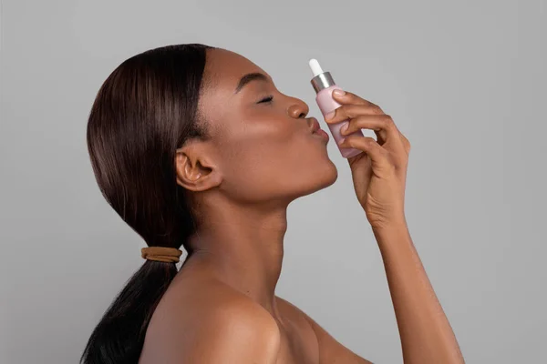 Feliz mulher negra bonita com perfeita pele pura beijando frasco de óleo, isolado em fundo cinza, perfil — Fotografia de Stock