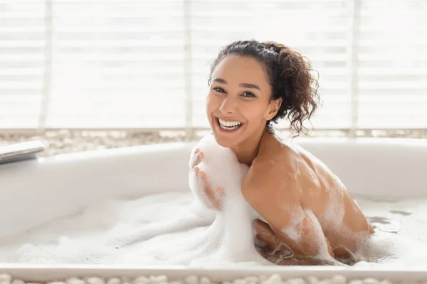 Charming joyful young woman sitting in hot bubble bath covered in foam, relaxing in bathtub at home, free space — ストック写真