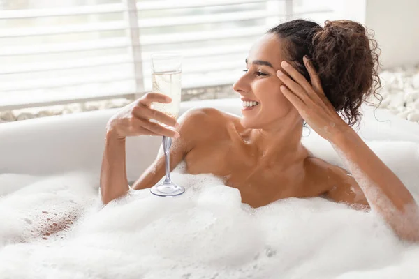Szczęśliwa młoda kobieta ciesząca się kieliszkiem szampana podczas relaksu w piankowej wannie w domu, puste miejsce — Zdjęcie stockowe