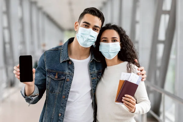Pareja joven con máscaras médicas sosteniendo Smartphone en blanco y pasaportes en el aeropuerto —  Fotos de Stock