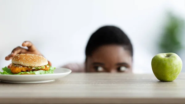 Mulher negra com excesso de peso olhando para hambúrguer de baixo da mesa, selecionando entre alimentos saudáveis e não saudáveis em casa — Fotografia de Stock