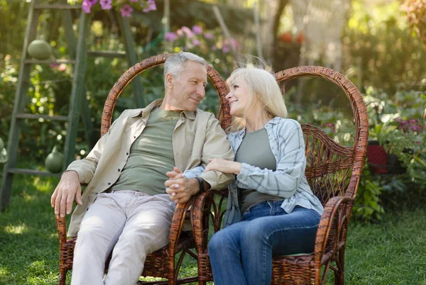 Šťastný starší manželé odpočívají venku na zahradě, sedí na proutěných židlích a dívají se na sebe — Stock fotografie
