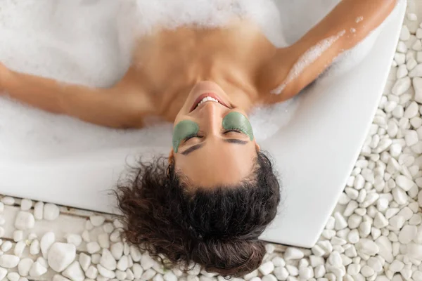 Pandangan utama wanita muda yang cantik santai dalam mandi busa dengan tambalan mata, menikmati prosedur perawatan kulit di rumah — Stok Foto