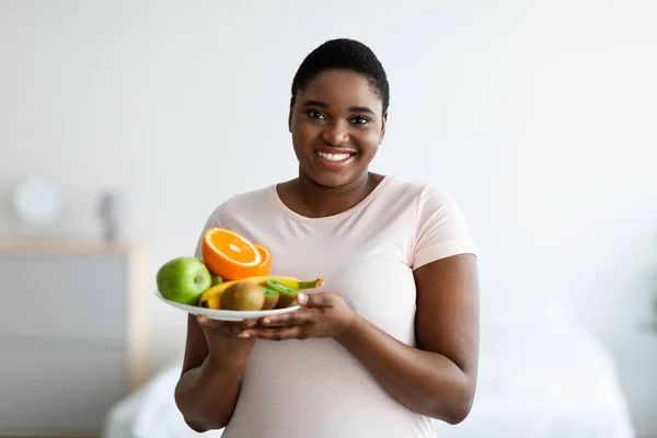 Sonriente mujer afroamericana con sobrepeso sosteniendo un plato de frutas frescas, manteniendo una dieta saludable para perder peso en casa —  Fotos de Stock