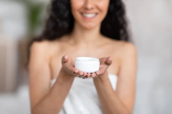 Smiling young caucasian brunette female with long hair in towel show jar of cream in bedroom interior, blurred