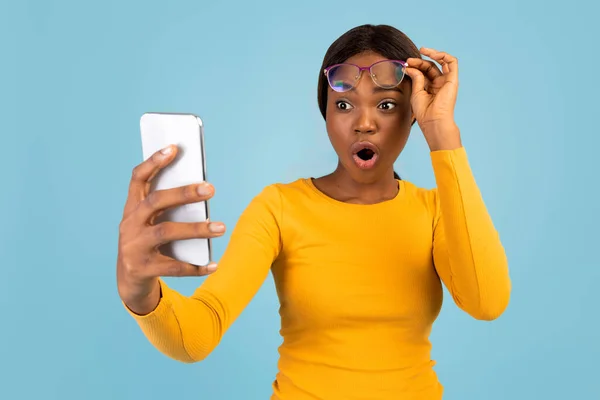 Megdöbbent fiatal fekete nő sárga pólóban leveszi a szemüveget, és néz okostelefon olvassa csodálatos üzenet — Stock Fotó