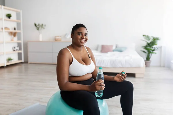 Además de tamaño joven mujer negra sentada en la pelota de fitness con botella de agua, haciendo ejercicio en casa durante el bloqueo covid —  Fotos de Stock