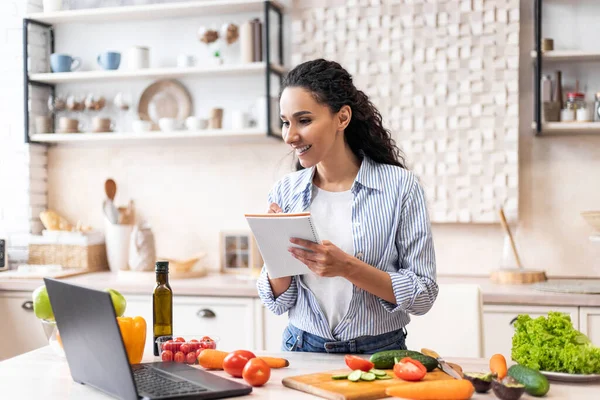 Positive Frau schreibt neues Rezept, während sie Video auf Laptop anschaut, Kochen in moderner Kücheneinrichtung — Stockfoto