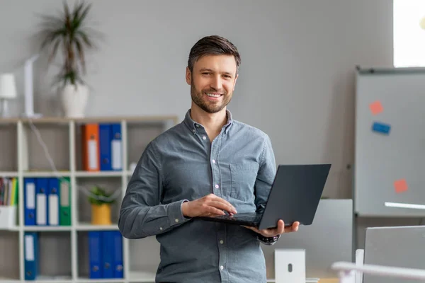 Porträtt av stilig mogen manlig kontorsarbetare med laptop i händerna stående på kontoret och ler mot kameran — Stockfoto