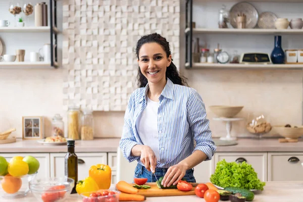 Vařím doma. Šťastná latinská žena připravuje zeleninový salát v moderní kuchyni, krájí okurku a usmívá se na kameru — Stock fotografie