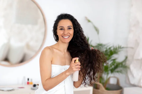 Lächelnde junge kaukasische brünette Dame im Handtuch sprüht Öl auf lange lockige Haare zur Behandlung im Schlafzimmer — Stockfoto