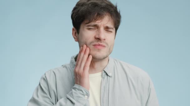 口腔衛生の問題。歯の痛みに苦しむ若い男痛みを伴う頬と悲しみ、青の背景に触れる — ストック動画