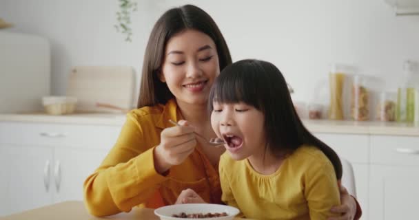 Ранкова рутина вдома. Симпатична любляча азіатська мати годує свою дочку молоком і крупами, сидячи на кухні — стокове відео