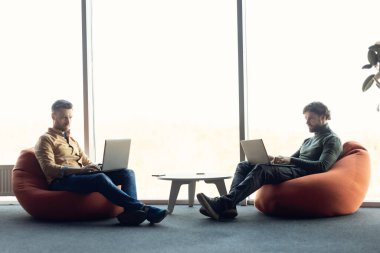 Fasulye çuvalındaki koltuklarda oturan, açık uzay ofisindeki dizüstü bilgisayarlarla çalışan, uzayı kopyalayan odaklanmış meslektaşlar.