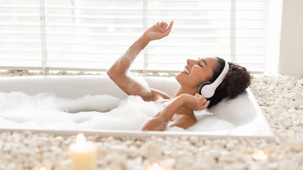 Позитивная юная леди в наушниках принимает пенную ванну, слушает музыку и поет любимую песню дома, баннер — стоковое фото