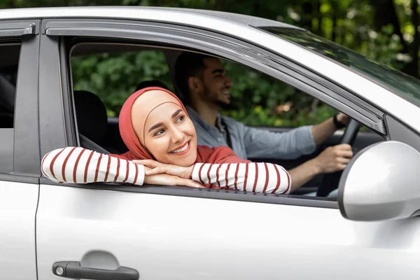 Glimlachende duizendjarige arabische vrouw in hijab rit in de auto kijkt uit het open raam buiten, genieten van reis — Stockfoto