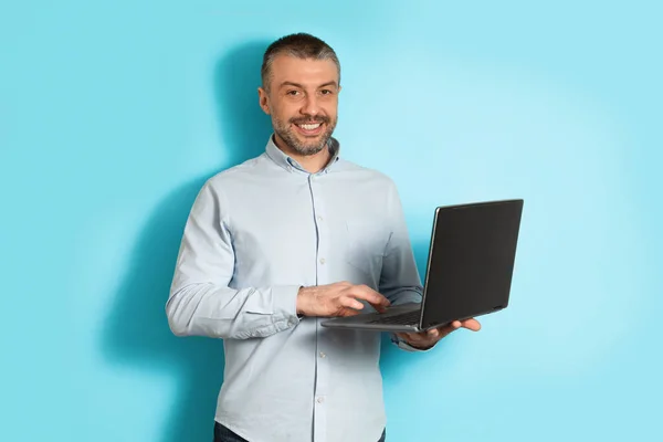 Homem de meia-idade usando computador portátil em pé sobre fundo azul — Fotografia de Stock