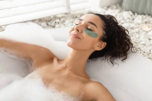 Charmante jeune femme allongée dans un bain moussant avec des taches sous les yeux, prenant soin de la peau délicate à la maison — Photo