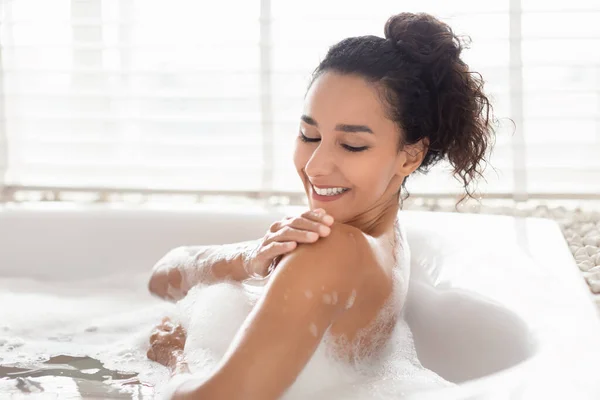 Mulher milenar encantadora deitada na banheira com espuma, desfrutando de banho de espuma, tocando pele sedosa macia no hotel, espaço livre — Fotografia de Stock