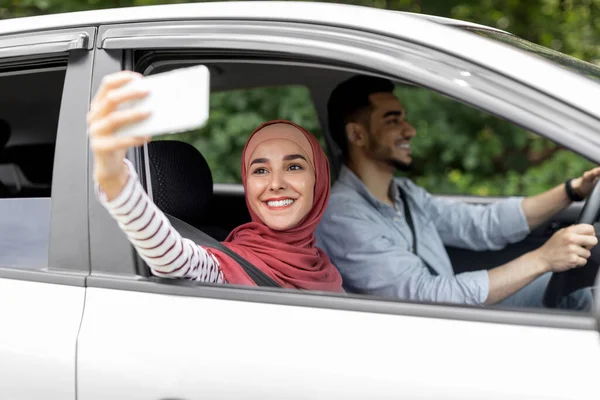 Blij millennial arabische vrouw in hijab neemt selfie of livestream op smartphone in open venster in de auto — Stockfoto