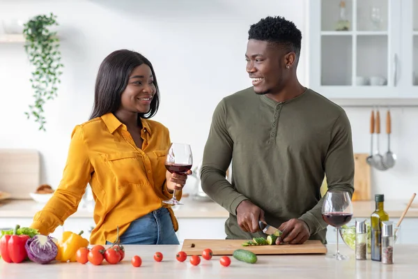 Feliz mujer negra bebiendo vino mientras su marido cocina ensalada en la cocina — Foto de Stock
