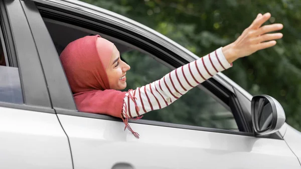 Tevreden duizendjarige islamitische vrouw in hijab vangt lucht met haar hand uit open raam in de auto, buiten, panorama — Stockfoto