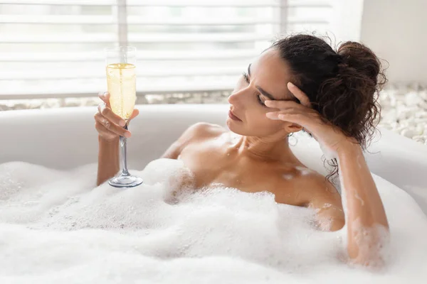 Mujer joven infeliz acostada en baño de burbujas con copa de champán en casa, espacio para copiar —  Fotos de Stock