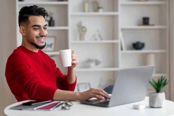 Wesoły, atrakcyjny młody arabski biznesmen z brodą w czerwonym ubraniu pije kawę i typy na laptopie w miejscu pracy — Zdjęcie stockowe