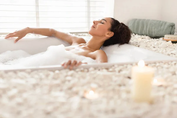 Belle jeune femme couchée dans un bain mousseux dans une atmosphère relaxante avec des bougies à la maison, espace libre — Photo