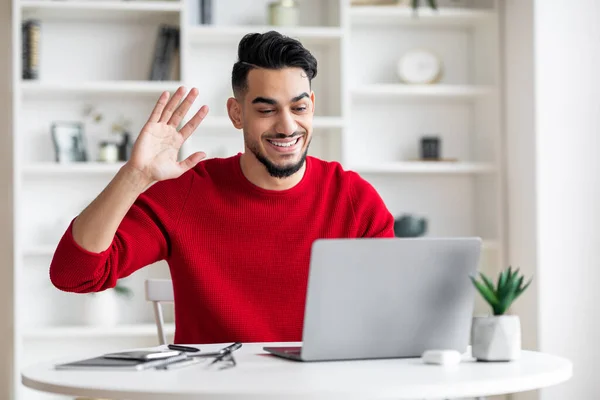 Feliz joven hombre islámico atractivo con barba en ropa roja agitando la mano, mira el ordenador portátil en el interior de la oficina en casa — Foto de Stock