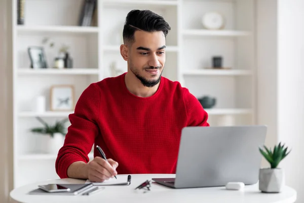 Lächelnd macht sich der attraktive Millennial-Araber mit Bart in roter Kleidung Notizen, arbeitet am Laptop — Stockfoto