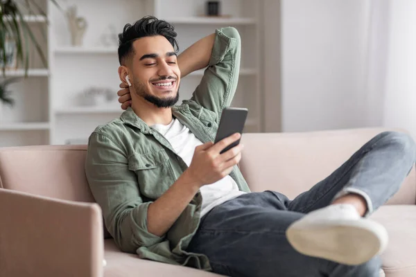 Szczęśliwy młody muzułmanin mężczyzna z brodą w przypadkowych, bezprzewodowych słuchawek siedzi na kanapie ze smartfonem i cieszy się ulubioną muzyką — Zdjęcie stockowe