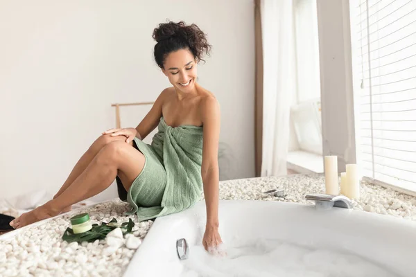 Charmante jonge vrouw in de handdoek maken schuimig bad, aanraken van water, temperatuur controleren, klaar voor thuis spa procedure — Stockfoto