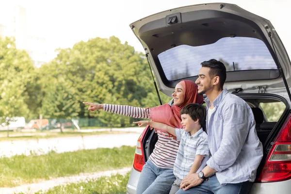 陽気な中東の妻でヒジャーブ,夫と小さな息子ポイント指に向かって空のスペース,車のトランクに座る — ストック写真