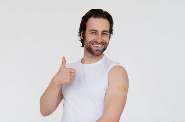 Happy young european man with stubble in t-shirt shows shoulder after vaccination and thumb up — Stock Photo, Image
