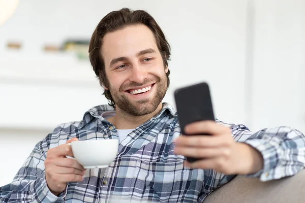 Sonriente joven europeo chico con rastrojo en camisa beber bebida caliente favorita y charlar en línea en el teléfono inteligente —  Fotos de Stock