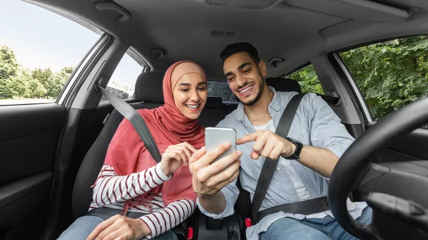 Lachende jonge arabische vrouw in hijab en man op zoek naar een manier op de kaart op de smartphone tijdens het rijden in de auto, buiten — Stockfoto