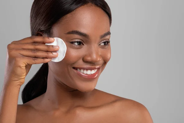 Mulher preta bonita feliz com pele perfeita removendo maquiagem com almofada de algodão isolada em fundo cinza — Fotografia de Stock