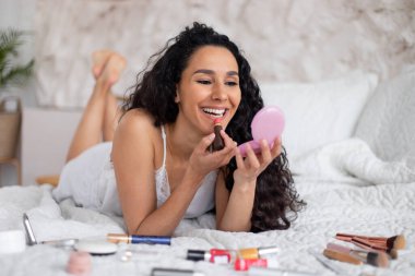 Uzun kıvırcık saçlı, pijamalı neşeli beyaz kadın yatağa uzanıyor ve dudaklarına ruj sürüyor.