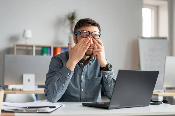 Зрелый работник-мужчина трёт усталые раздражённые глаза, слишком много работает с ноутбуком, устал от онлайн-работы в офисе — стоковое фото