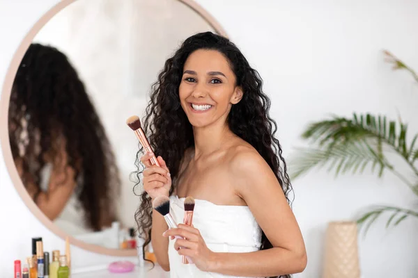 Lächelnde junge kaukasische brünette Dame mit langen lockigen Haaren im Handtuch mit Make-up-Pinseln in der Nähe des Spiegels — Stockfoto